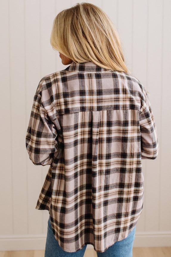 A woman viewed from behind wearing a long-sleeve plaid button-up shirt in beige, brown, and black, paired with blue jeans against a white paneled wall.