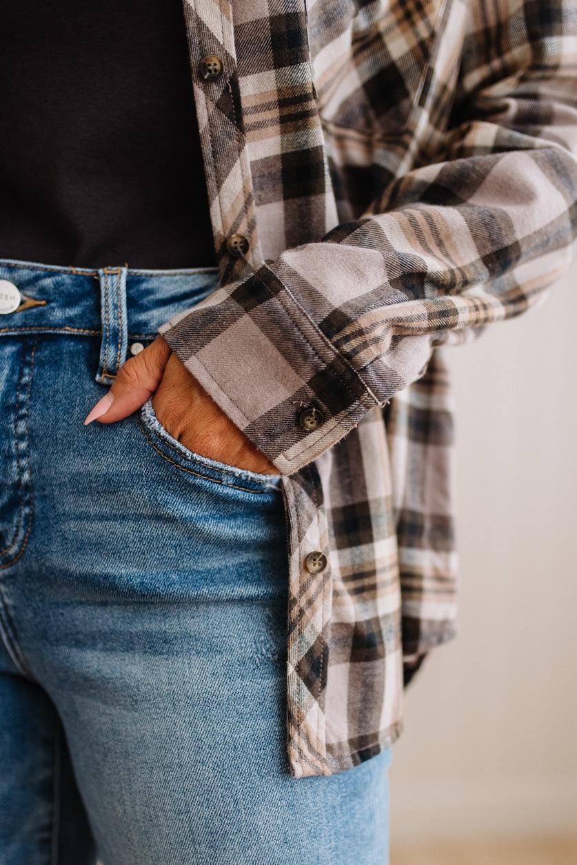 A close-up of a person wearing a long-sleeve plaid button-up shirt in beige, brown, and black over a black top, paired with blue jeans.