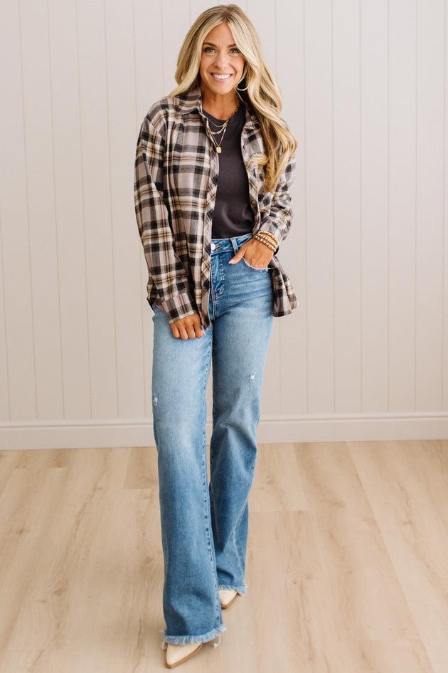 A woman modeling a brown and beige plaid button-down shirt over a black top, paired with light-wash blue flare jeans and beige pointed-toe heels, standing in a room with light wood floors and white paneled walls.
