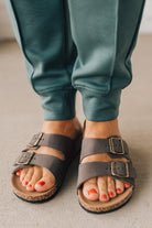 Woman wearing ash jade joggers zoomed in to show brown slip on sandals with double strap and brushed gold buckles.