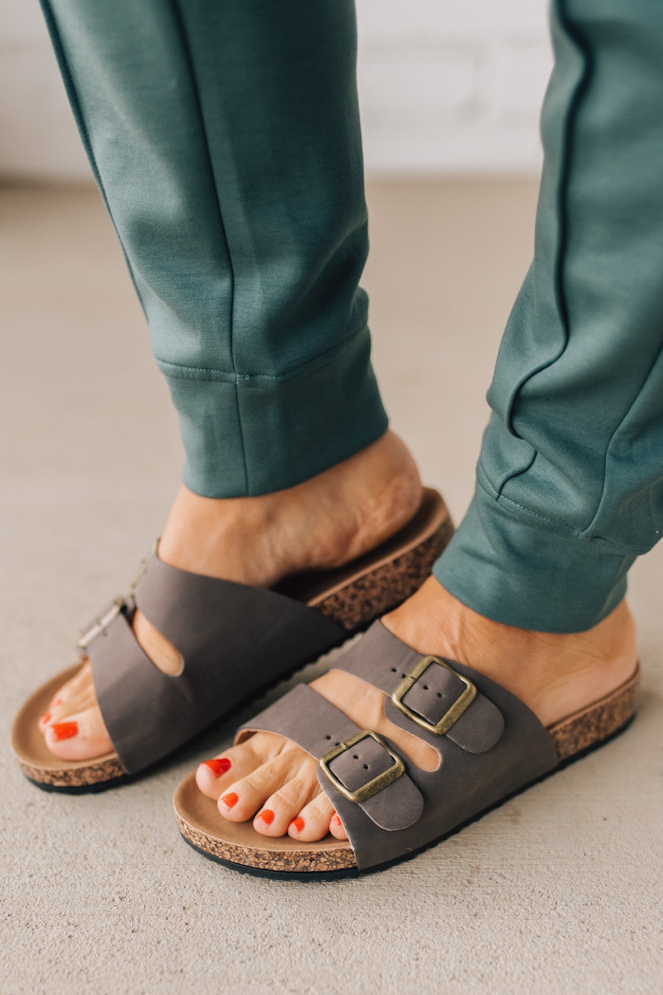 Woman wearing ash jade joggers zoomed in to show brown slip on sandals with double strap and brushed gold buckles.