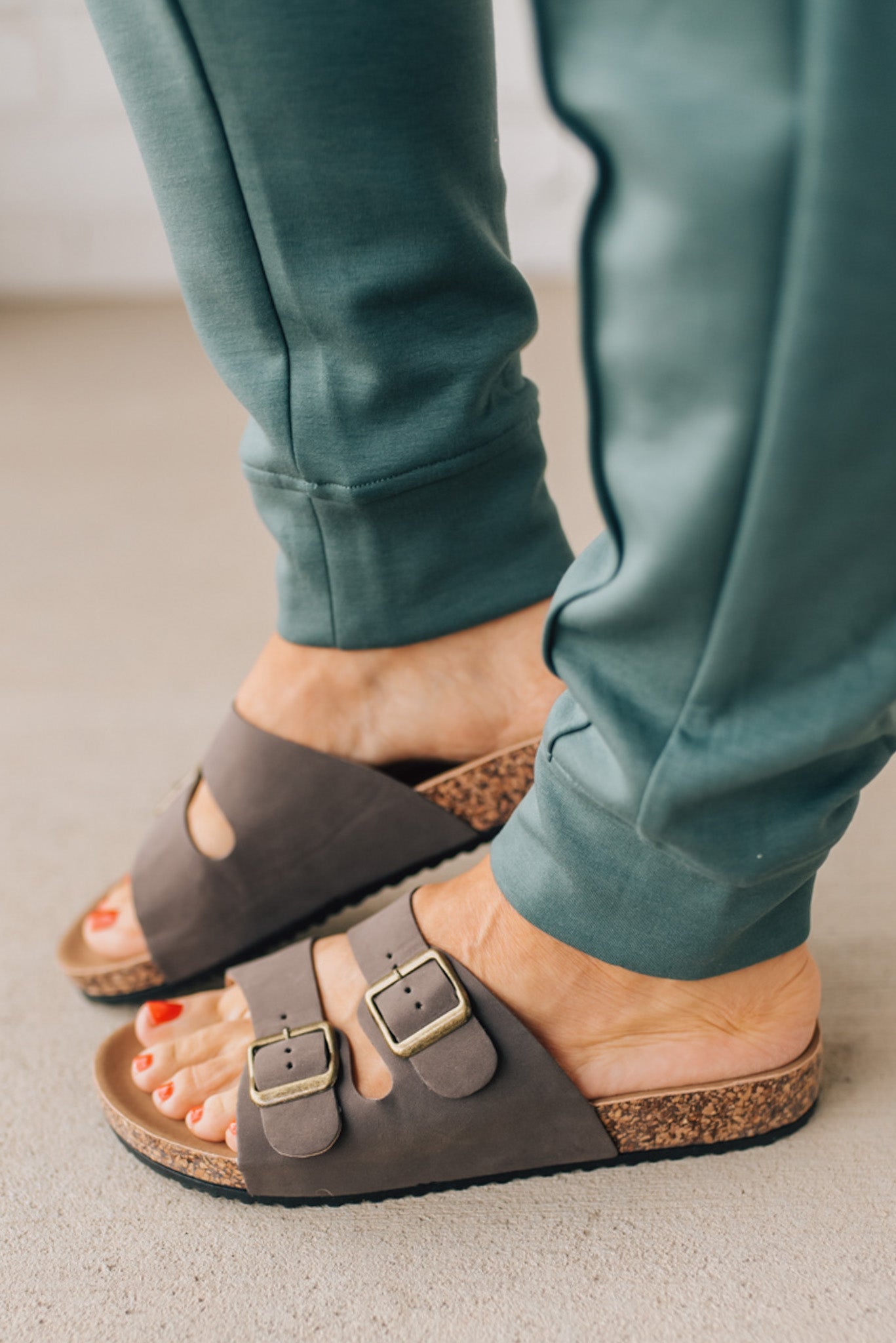 Woman wearing ash jade joggers zoomed in to show brown slip on sandals with double strap and brushed gold buckles.