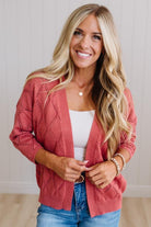 A smiling blonde woman wearing a dusty rose cable-knit cardigan over a white tank top and blue jeans, standing against a light wood-paneled wall.