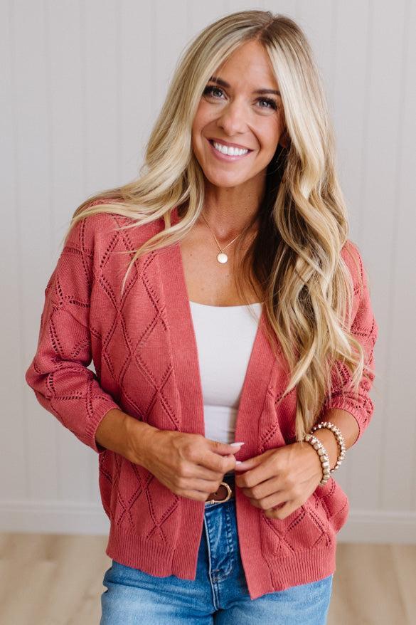 A smiling blonde woman wearing a dusty rose cable-knit cardigan over a white tank top and blue jeans, standing against a light wood-paneled wall.
