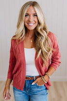 A smiling blonde woman wearing a dusty rose cable-knit cardigan over a white tank top and blue jeans, standing against a light neutral wall.