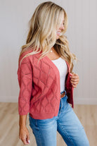 A woman wearing a coral pink open-front knit cardigan with a diamond lattice pattern over a white top and blue jeans, standing in a brightly lit indoor setting.