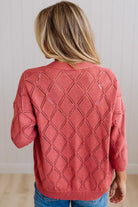 A woman viewed from behind wearing a coral pink knit sweater with an openwork diamond pattern, paired with blue jeans, set against a neutral indoor background.