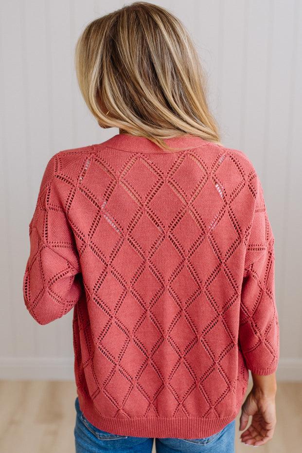 A woman viewed from behind wearing a coral pink knit sweater with an openwork diamond pattern, paired with blue jeans, set against a neutral indoor background.