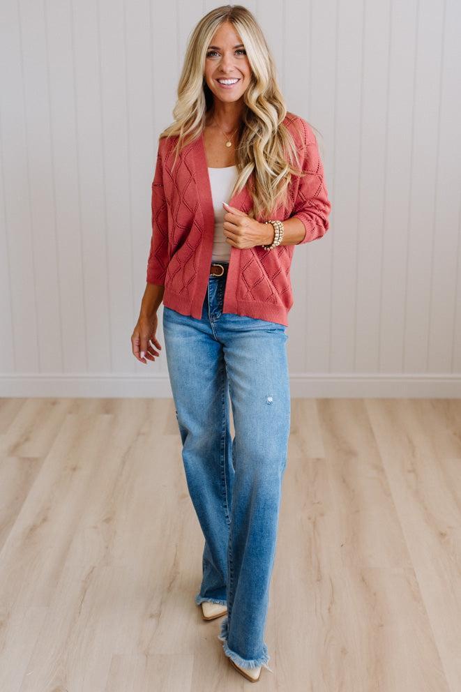 A woman modeling a dusty rose quilted jacket, blue jeans, and a white top in a bright room with light wood floors and white paneled walls.