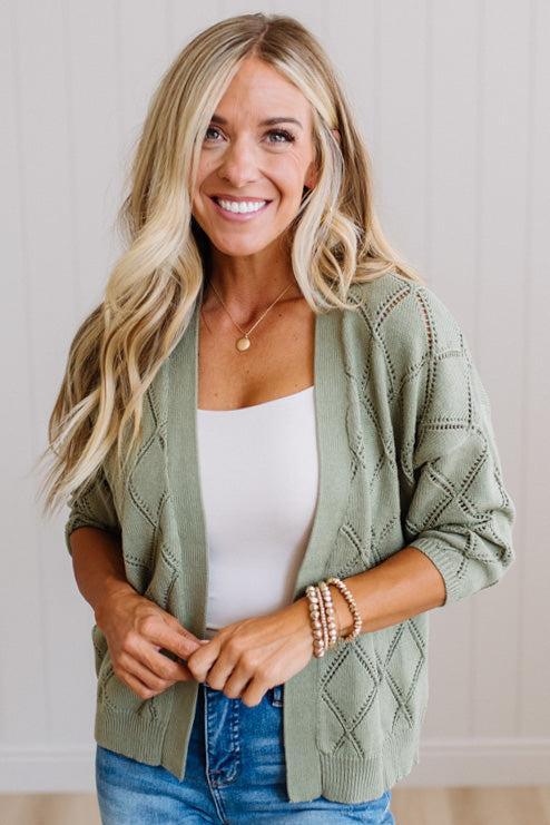 A smiling blonde woman wearing a sage green cable-knit cardigan over a white tank top and blue jeans, standing against a light paneled wall.