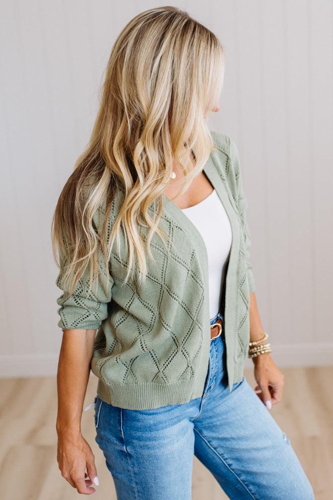 A woman wearing a sage green knit cardigan with a diamond pattern over a white top and blue jeans, standing in a brightly lit room with light wood floors and white paneled walls.