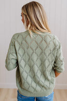 A woman viewed from behind wearing a sage green knit sweater with a textured diamond lattice pattern, paired with blue jeans, set against a light wood-paneled wall.