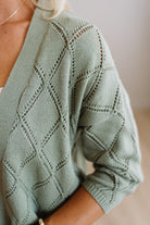 A close-up of a person wearing a sage green knit cardigan with a textured diamond lattice pattern and openwork details.