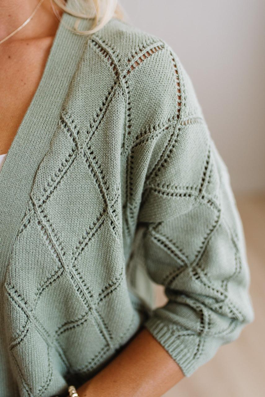 A close-up of a person wearing a sage green knit cardigan with a textured diamond lattice pattern and openwork details.
