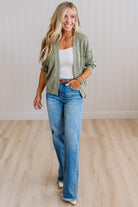 A woman modeling a sage green cable-knit cardigan over a white tank top and blue jeans, standing in a room with light wood floors and white paneled walls.