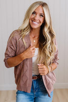 A smiling blonde woman wearing a dusty rose knit cardigan over a white tank top and blue jeans, standing against a light paneled wall.