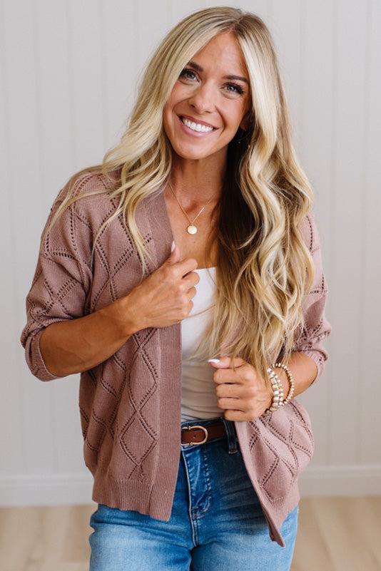 A smiling blonde woman wearing a dusty rose knit cardigan over a white tank top and blue jeans, standing against a light paneled wall.