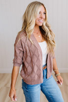 A smiling blonde woman wearing a dusty rose open-front knit cardigan with a diamond lattice pattern over a white tank top and blue jeans, standing against a light neutral wall.