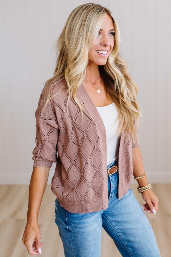 A smiling blonde woman wearing a dusty rose open-front knit cardigan with a diamond lattice pattern over a white tank top and blue jeans, standing against a light neutral wall.