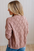 A woman shown from the back wearing a dusty rose pink knit cardigan with a diamond lattice pattern, paired with blue jeans, set against a light-colored paneled wall.