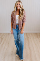 A woman stands in a bright room with light wood flooring and white paneled walls, wearing a dusty rose knit cardigan over a white tank top, blue jeans, and beige heels.