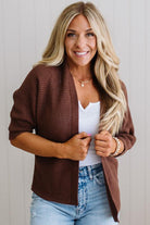 Woman wearing a brown cardigan over a white top and blue jeans, standing against a plain background.