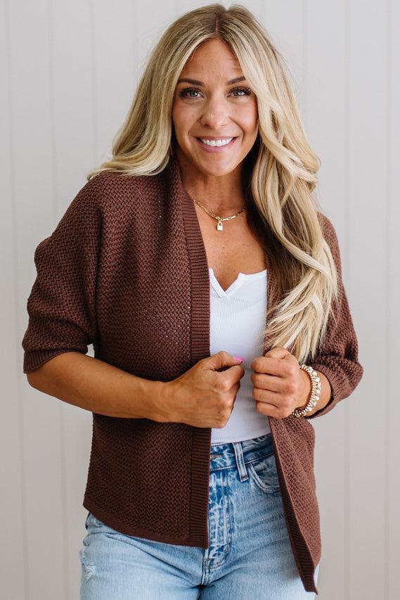 Woman wearing a brown cardigan over a white top and blue jeans, standing against a plain background.