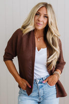 Woman wearing a brown cardigan over a white top and blue jeans, standing against a plain background.