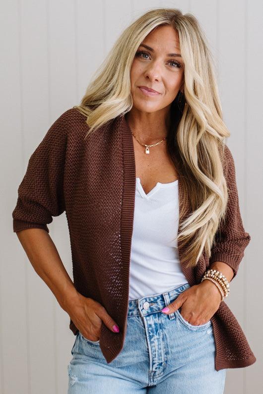 Woman wearing a brown cardigan over a white top and blue jeans, standing against a plain background.