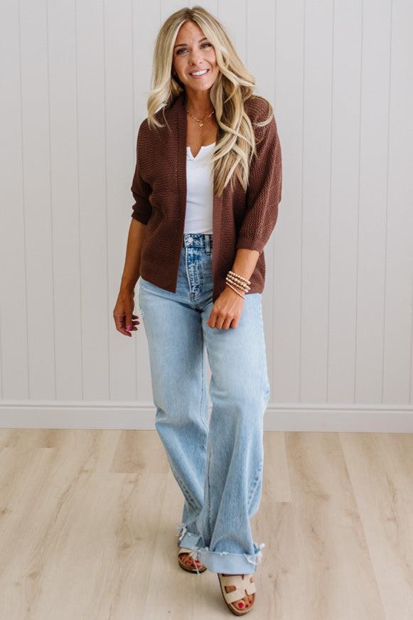 Woman wearing a brown cardigan, white top, and light blue jeans indoors.