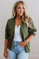 Woman wearing a green cardigan over a white top and blue jeans, standing against a light wooden panel background.