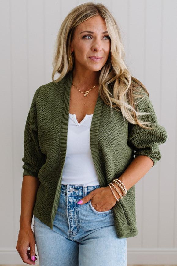 Woman wearing a green cardigan over a white top and blue jeans, standing against a light wooden panel background.