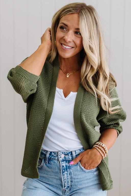 Woman wearing a green cardigan over a white shirt and blue jeans, smiling.