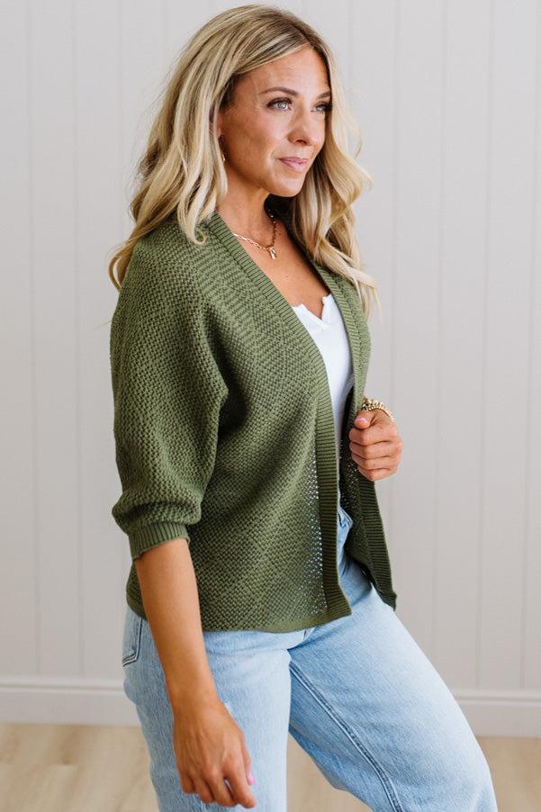 Woman wearing a green cardigan over a white top and light blue jeans, sitting against a plain background.
