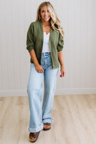 Woman wearing a green cardigan, white top, and light blue jeans against a white wall.