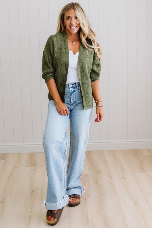 Woman wearing a green cardigan, white top, and light blue jeans against a white wall.