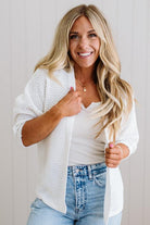 Woman wearing a white cardigan over a white shirt and blue jeans, smiling against a plain background