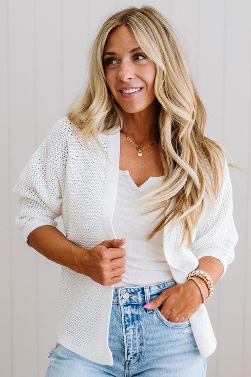 Woman wearing a white cardigan over a white top and blue jeans, standing against a light background.