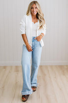 Woman wearing a white blazer, white shirt, and light blue jeans standing in a room with a white wall and wooden floor.