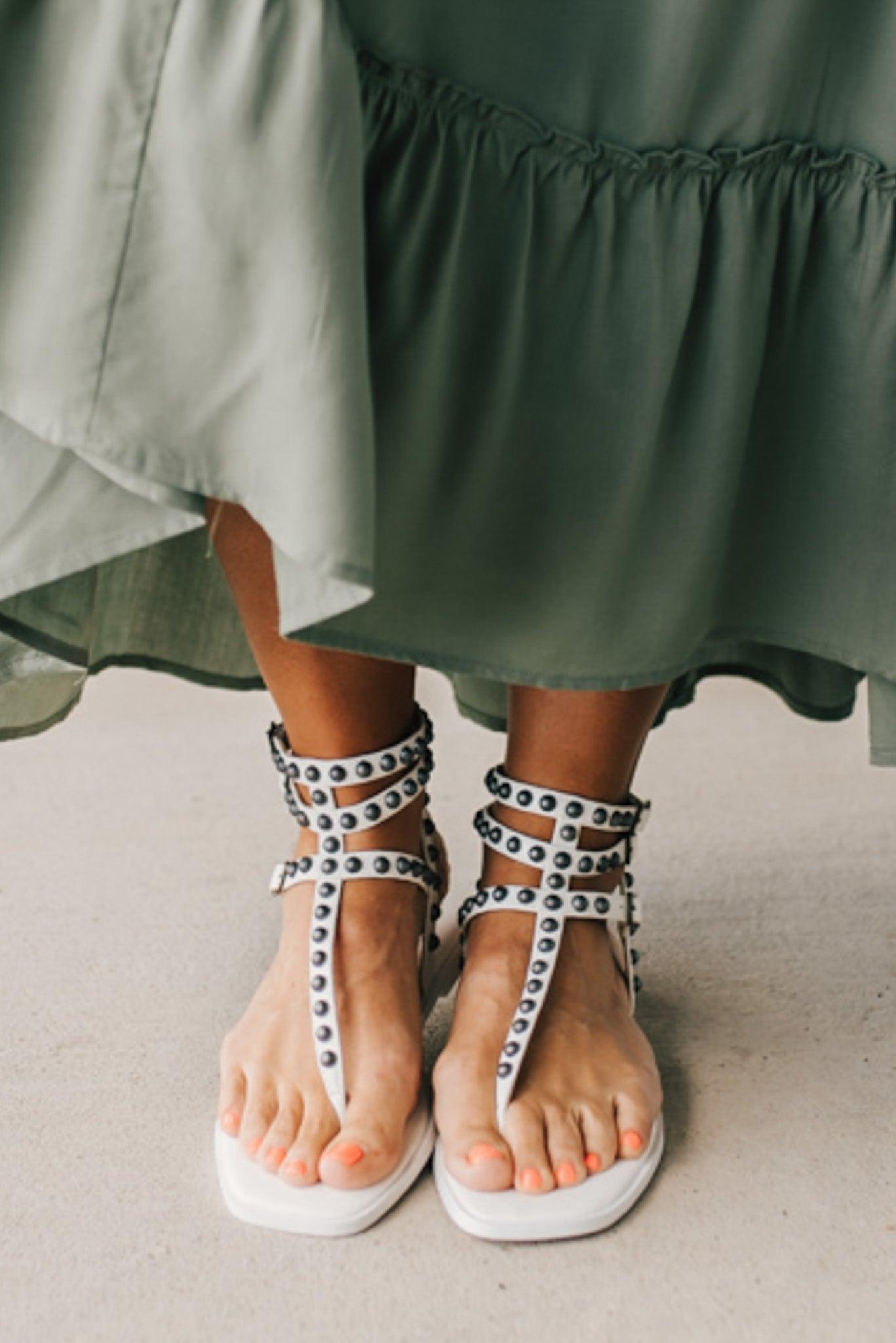 Woman in a green skirt wearing flat white faux leather gladiator sandals t-strap with triple buckle around ankle and pewter stud accents on straps.