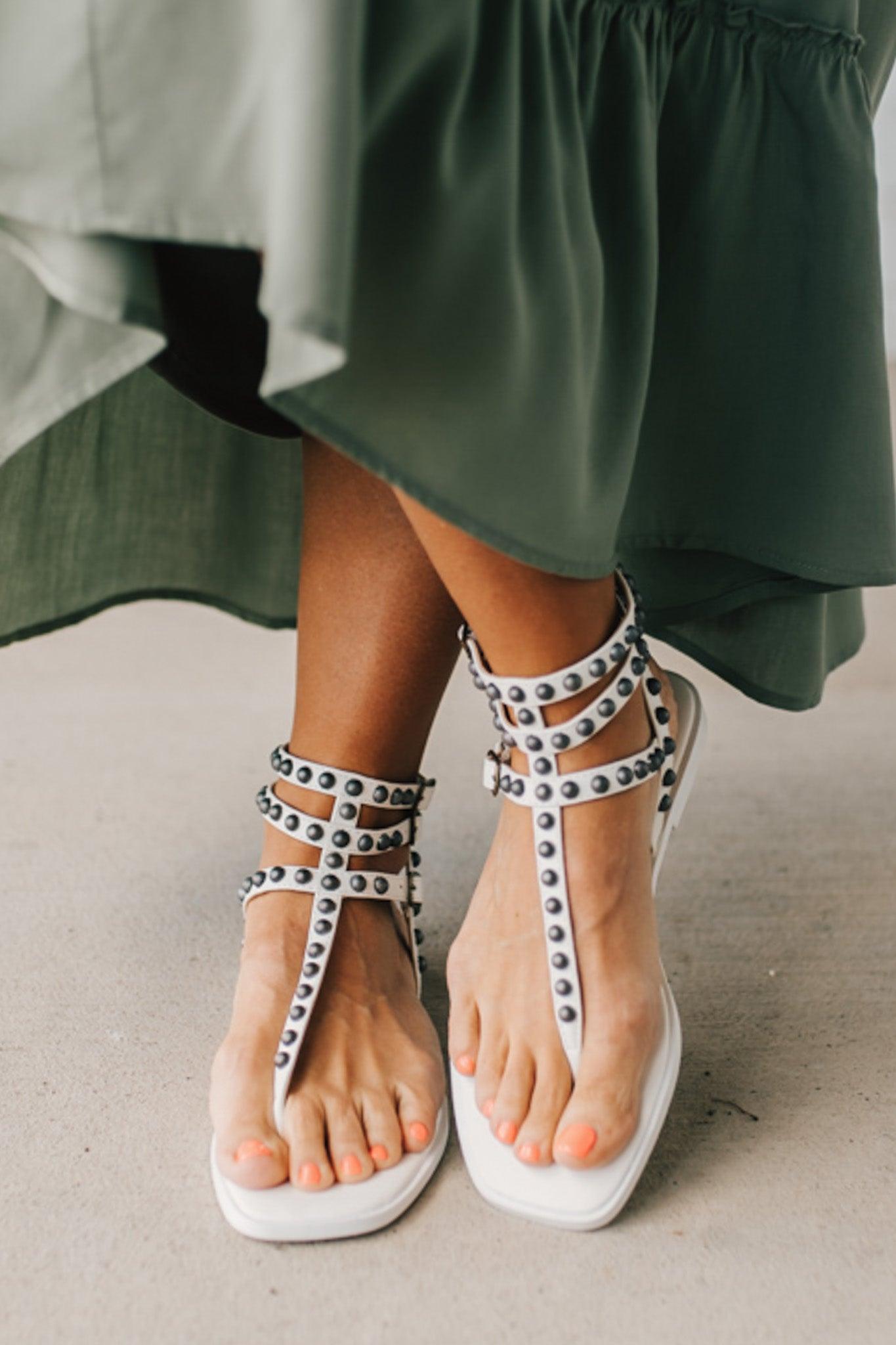 Woman in a green skirt wearing flat white faux leather gladiator sandals t-strap with triple buckle around ankle and pewter stud accents on straps.