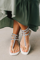 Woman in a green skirt wearing flat white faux leather gladiator sandals t-strap with triple buckle around ankle and pewter stud accents on straps.
