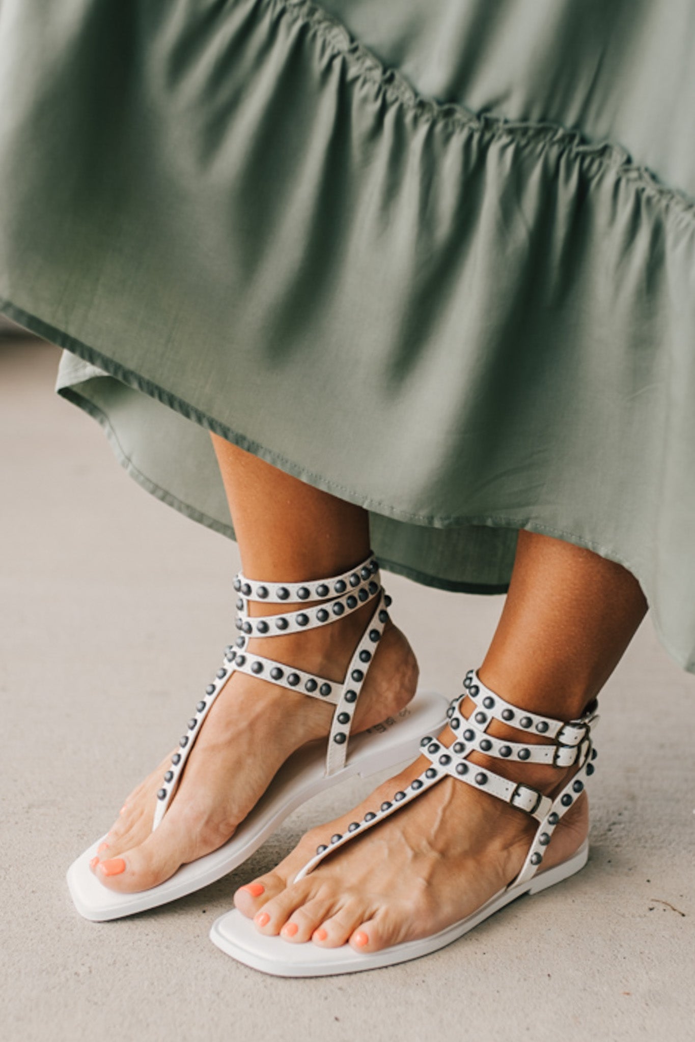 Woman in a green skirt wearing flat white faux leather gladiator sandals t-strap with triple buckle around ankle and pewter stud accents on straps.