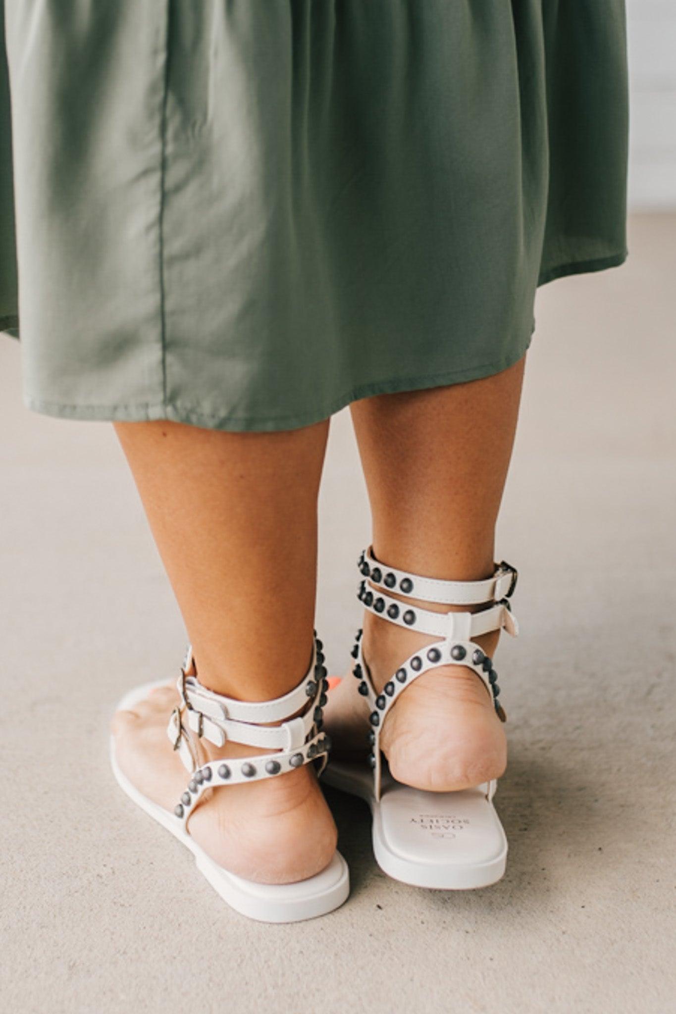 Woman in a green skirt wearing flat white faux leather gladiator sandals t-strap with triple buckle around ankle and pewter stud accents on straps.
