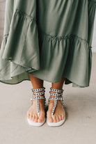 Woman in a green skirt wearing flat white faux leather gladiator sandals t-strap with triple buckle around ankle and pewter stud accents on straps.