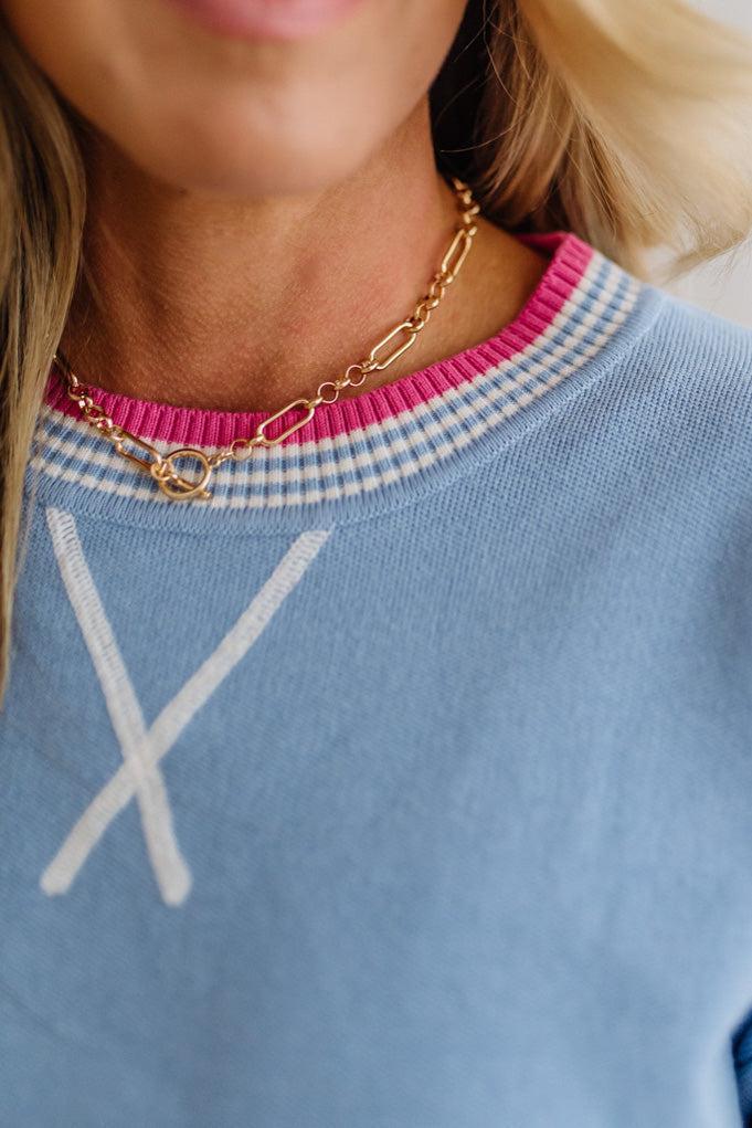 Close-up of a person wearing a blue sweater with a pink and white striped collar and a gold necklace.