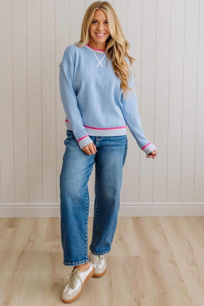 Woman wearing a light blue sweater with pink accents and jeans, standing against a white paneled wall.
