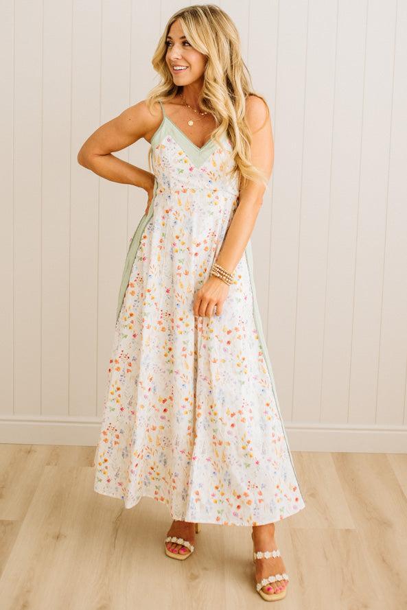 Woman wearing a floral dress standing against a light wooden paneled wall.