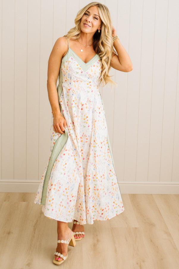 Woman wearing a floral dress holding a green bag against a light wooden paneled wall.