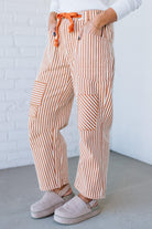 Person wearing brown and white striped pants with orange drawstring against a white brick wall.